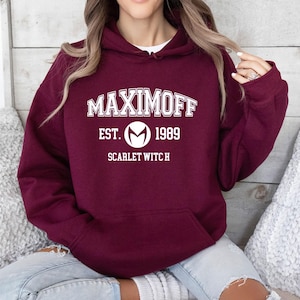 Op de afbeelding: Bordeaux hoodie met een voorzak en een witte afbeelding. De afbeelding luidt "MAXIMOFF EST. 1989 SCARLET WITCH" in een vetgedrukte lettertype in collegestijl. Het ontwerp bevat een cirkelvormig embleem. De hoodie is gemaakt van zacht materiaal.
