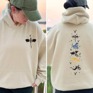 May include: Beige hoodie with a front pocket and a dragon graphic. The front features a dragon with a crescent moon and stars. The back displays a vertical arrangement of dragon illustrations in various colors, including black, blue, and yellow.