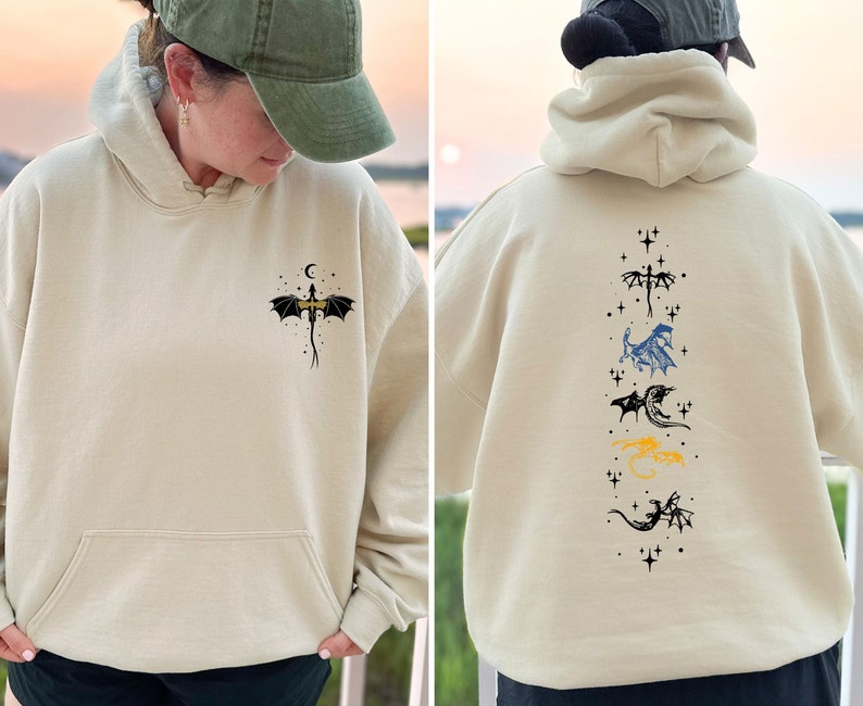 May include: Beige hoodie with a front pocket and a dragon graphic. The front features a dragon with a crescent moon and stars. The back displays a vertical arrangement of dragon illustrations in various colors, including black, blue, and yellow.