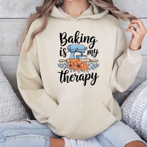 Puede incluir: Sudadera con capucha beige con bolsillo delantero y cordón. El gráfico en la parte delantera dice "Baking is my therapy" con una batidora azul, un rodillo y flores naranjas y azules.