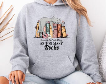 Puede incluir: Sudadera con capucha gris claro con un gráfico de un gato gris durmiendo sobre libros y flores. El texto dice "There Is No Such Thing As TOO MANY Books". La sudadera tiene un bolsillo delantero y una capucha.