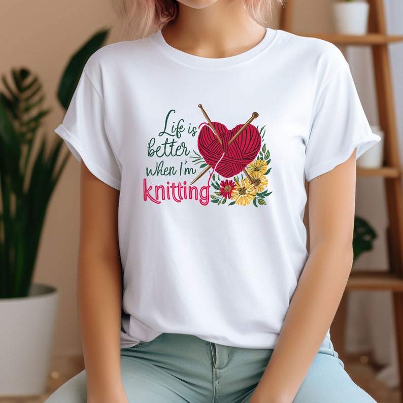 May include: White t-shirt with a graphic design of a red heart with yarn and knitting needles, surrounded by flowers. The text "Life is better when I'm knitting" is printed on the shirt.