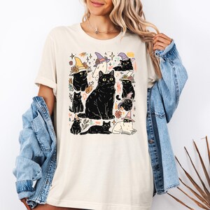 May include: A cream-colored t-shirt featuring a graphic print of multiple black cats, some wearing colorful pointed hats, interspersed with small floral and star illustrations. The design includes a large central cat surrounded by smaller cat figures.