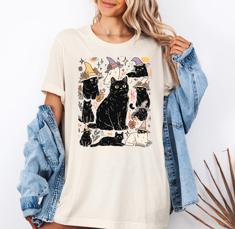 May include: A cream-colored t-shirt featuring a graphic print of multiple black cats, some wearing colorful pointed hats, interspersed with small floral and star illustrations. The design includes a large central cat surrounded by smaller cat figures.