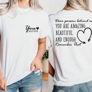 Op de afbeelding: Wit T-shirt met de tekst "You Matter" en een klein hartje op de voorkant. De achterkant van het shirt heeft de tekst "Dear person behind me, YOU ARE AMAZING BEAUTIFUL AND ENOUGH Remember That" met een hartje en een pijl.