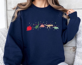 May include: Navy blue sweatshirt featuring a whimsical design of a red sleigh pulled by a line of colorful dinosaurs. The dinosaurs are green, pink, yellow, and green, with white star accents. A fun, festive design for the holiday season.