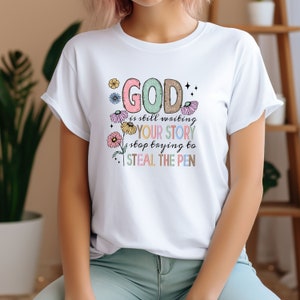 May include: White t-shirt with a colourful graphic design that says "GOD is still writing YOUR STORY stop trying to STEAL THE PEN".