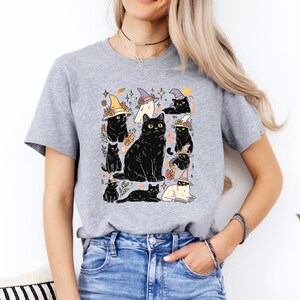 May include: A heather gray short-sleeve t-shirt featuring a graphic print of multiple black cats, some wearing colorful witch hats, interspersed with small floral elements, stars, and a ghost, all arranged in a central cluster on the chest.