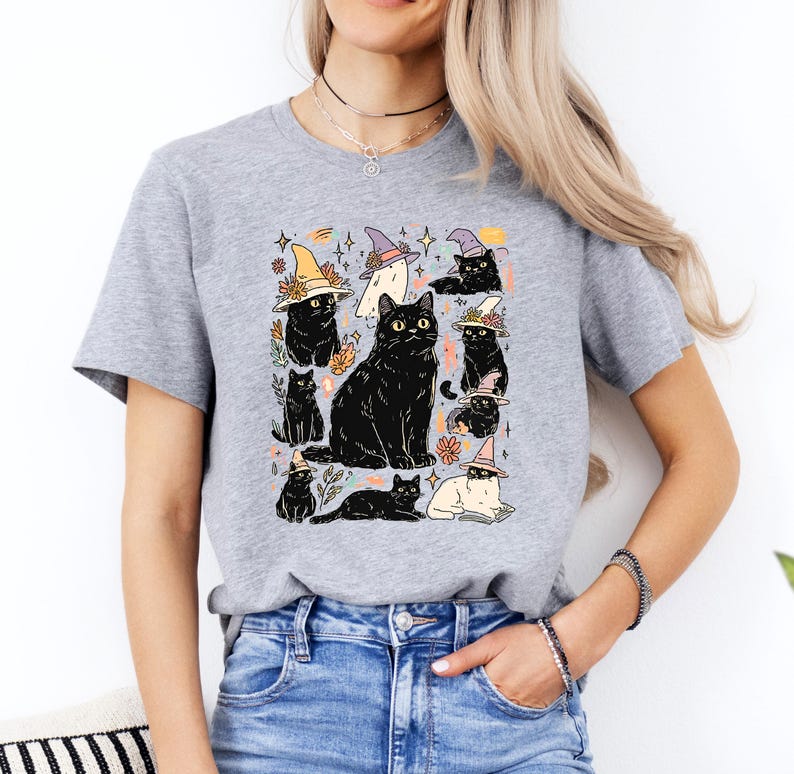 May include: A heather gray short-sleeve t-shirt featuring a graphic print of multiple black cats, some wearing colorful witch hats, interspersed with small floral elements, stars, and a ghost, all arranged in a central cluster on the chest.