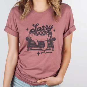 May include: A mauve t-shirt with a graphic of a cat sleeping on a couch. The text "Sorry got plans" is printed below the cat.