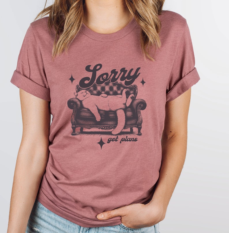 May include: A mauve t-shirt with a graphic of a cat sleeping on a couch. The text "Sorry got plans" is printed below the cat.