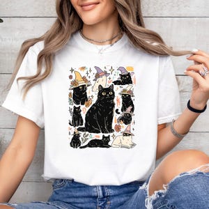 May include: A white short-sleeve t-shirt featuring a graphic print of multiple black cats wearing colorful witch hats. The illustration includes various cat poses, small floral accents, and star shapes scattered around the central cat figures.