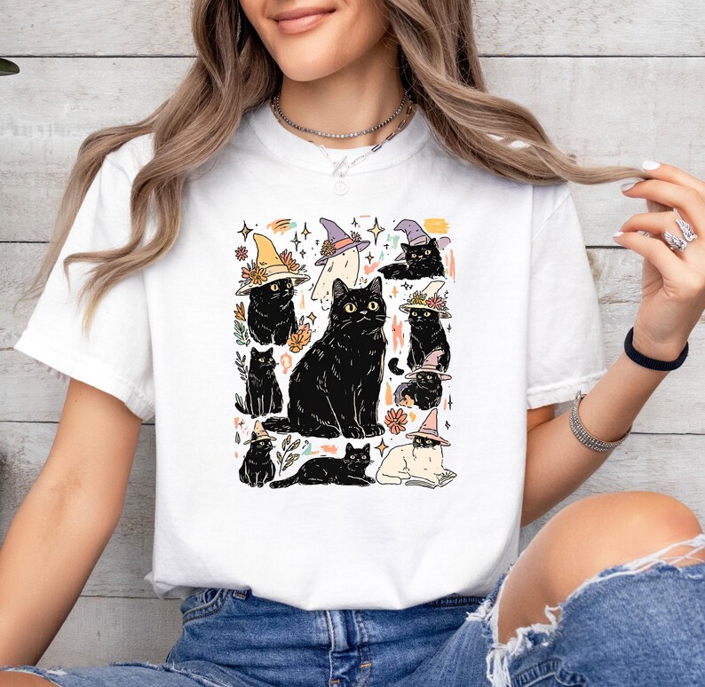 May include: A white short-sleeve t-shirt featuring a graphic print of multiple black cats wearing colorful witch hats. The illustration includes various cat poses, small floral accents, and star shapes scattered around the central cat figures.