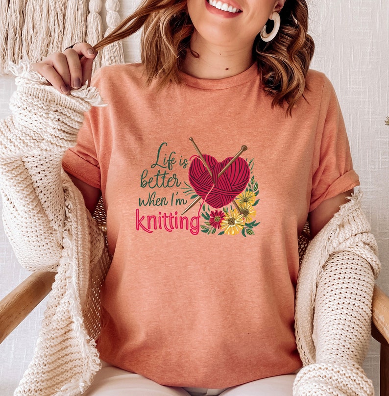 May include: A peach-colored t-shirt with the text "Life is better when I'm knitting" and a graphic of a heart-shaped ball of yarn with knitting needles and flowers.