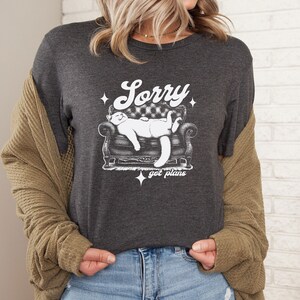 May include: A dark grey t-shirt with a white graphic of a cat sleeping on a couch. The text "Sorry got plans" is printed below the cat.