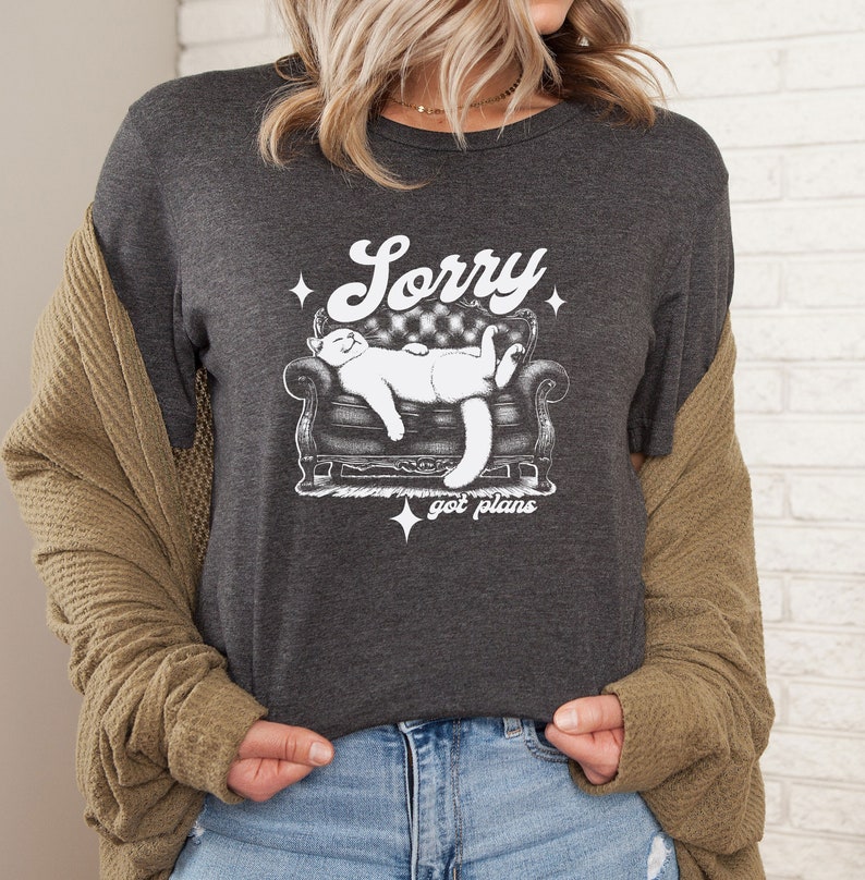 May include: A dark grey t-shirt with a white graphic of a cat sleeping on a couch. The text "Sorry got plans" is printed below the cat.