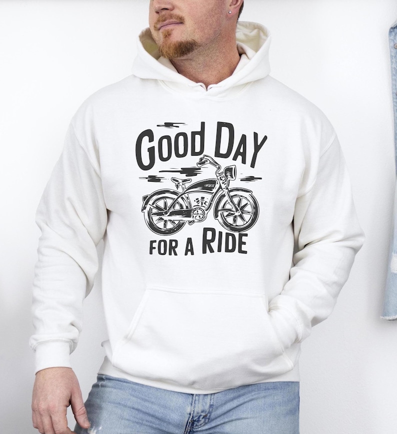 Może przedstawiać: Kremowa bluza z kapturem z czarnym nadrukiem graficznym przedstawiającym rower w stylu vintage na środku klatki piersiowej. Napis "GOOD DAY FOR A RIDE" jest wydrukowany pogrubioną, ciemną czcionką nad i pod ilustracją roweru z przodu.
