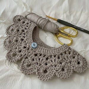 Puede incluir: Un cuello de ganchillo beige hecho a mano con borde festoneado y cierre de bot&oacute;n. Una madeja de hilo a juego, un ganchillo y unas tijeras doradas sugieren un tema artesanal.