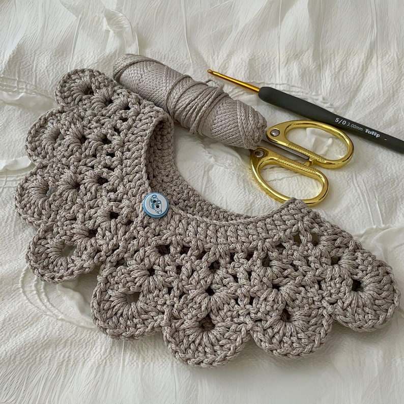 Puede incluir: Un cuello de ganchillo beige hecho a mano con borde festoneado y cierre de bot&oacute;n. Una madeja de hilo a juego, un ganchillo y unas tijeras doradas sugieren un tema artesanal.