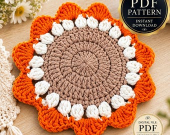 May include: A round crochet coaster featuring a brown center, a ring of white puff stitches, and an outer border of orange scalloped petals. The image includes a gold circular badge with the text PDF PATTERN INSTANT DOWNLOAD.