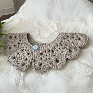 Puede incluir: Un cuello de crochet beige neutro, con un dise&ntilde;o de borde festoneado. El cuello se abrocha con un peque&ntilde;o bot&oacute;n redondo con una ilustraci&oacute;n azul y blanca. El patr&oacute;n de crochet calado es visible en todo el cuello.