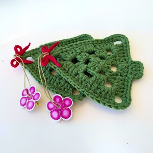 K&ouml;nnte beinhalten: Zwei gr&uuml;ne geh&auml;kelte Weihnachtsbaumschmuckst&uuml;cke mit roten Schleifen und rosa Blumenverzierungen.