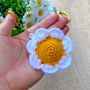 Puede incluir: Un llavero de margarita tejido a crochet con un centro amarillo y pétalos blancos. El llavero está unido a un anillo de llave dorado.