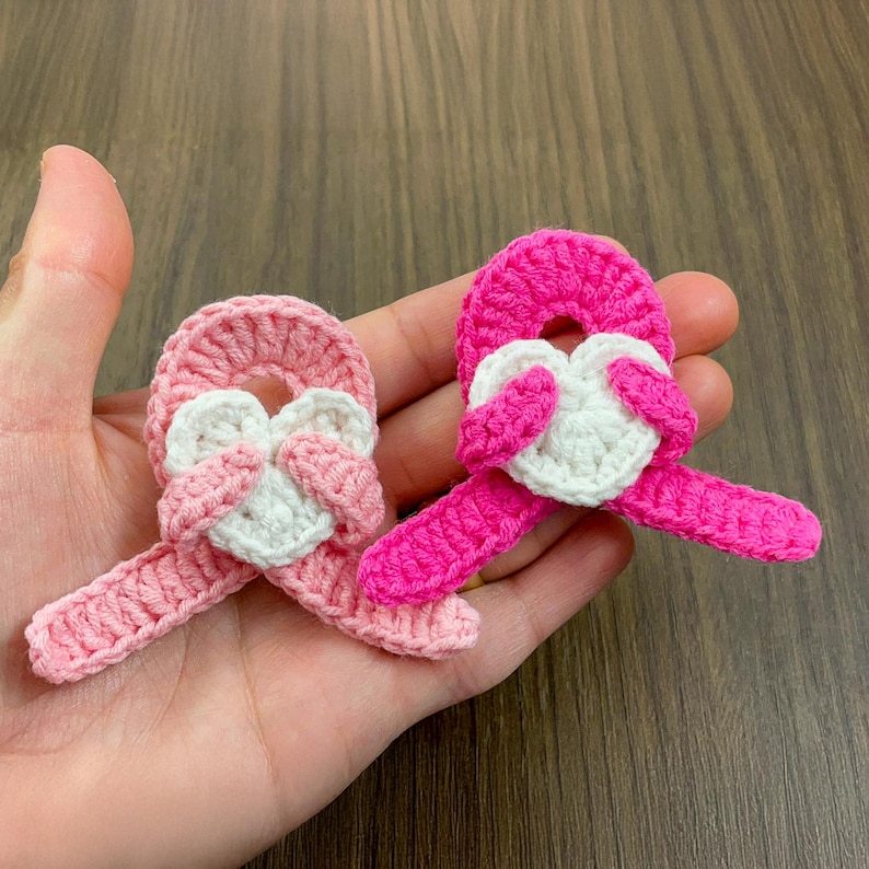 May include: Two handmade crochet ribbons in shades of pink and white. Each ribbon features a heart-shaped centerpiece and a looped design. The items are held in a hand, showcasing the texture and detail of the yarn.