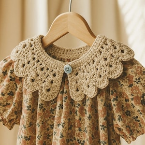 Puede incluir: Vestido con estampado floral y cuello de ganchillo. El vestido presenta un cuello de ganchillo color beige con borde festoneado y cierre de bot&oacute;n peque&ntilde;o. El vestido tiene mangas cortas y un estampado floral en tonos marrones y naranjas.
