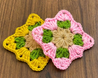 May include: Two crocheted squares, one pink and one yellow, with green accents. The squares are made with a granny stitch pattern.