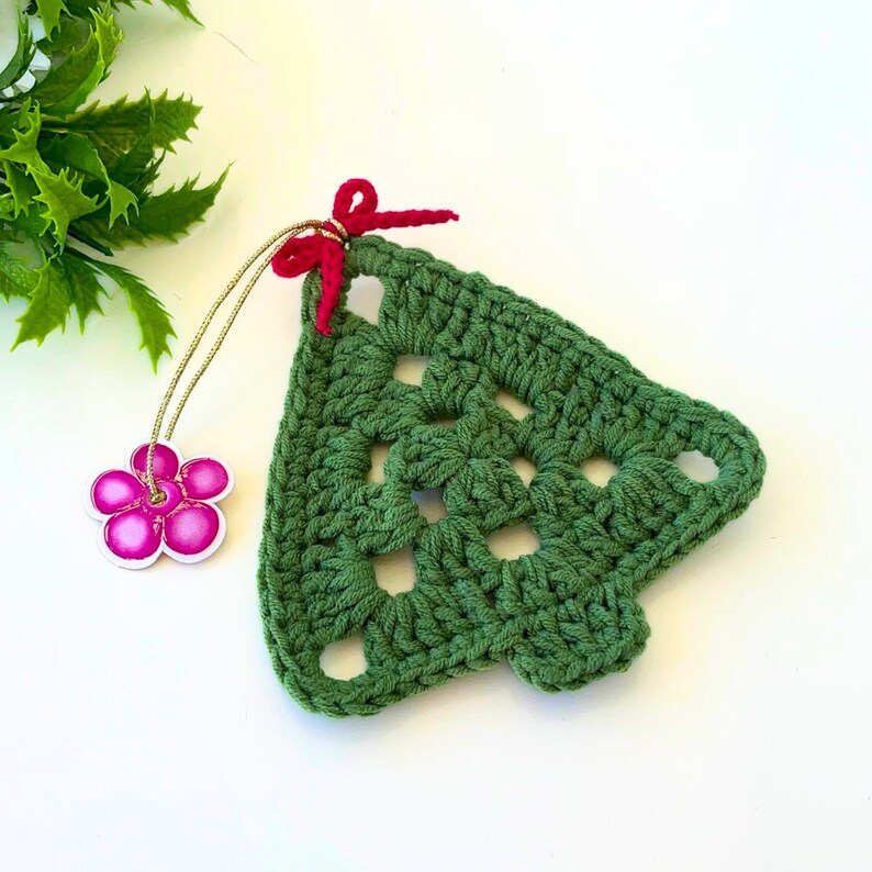 K&ouml;nnte beinhalten: Gr&uuml;ner geh&auml;kelter Weihnachtsbaum-Anh&auml;nger mit einer roten Schleife und einem rosa Blumenanh&auml;nger.