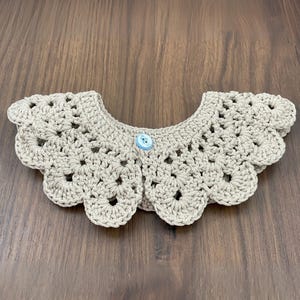 Puede incluir: Un cuello de ganchillo beige claro con un borde festoneado y un bot&oacute;n azul claro. El cuello presenta un dise&ntilde;o calado. El art&iacute;culo est&aacute; hecho de hilo.