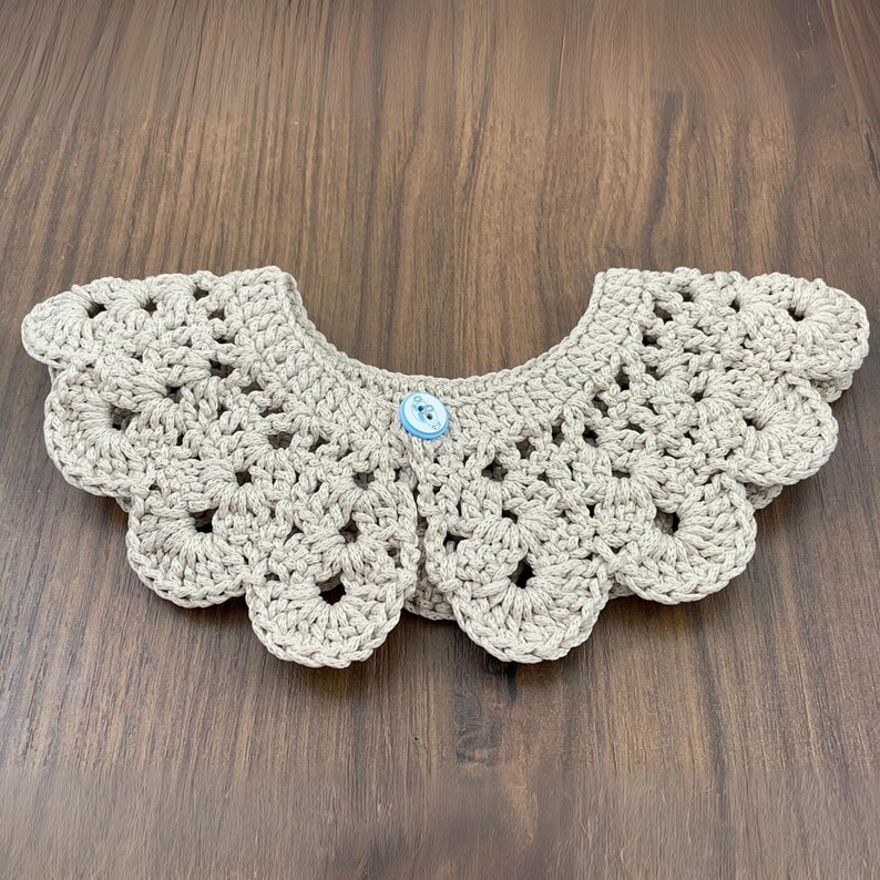 Puede incluir: Un cuello de ganchillo beige claro con un borde festoneado y un bot&oacute;n azul claro. El cuello presenta un dise&ntilde;o calado. El art&iacute;culo est&aacute; hecho de hilo.