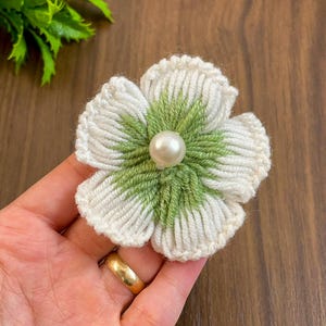 Puede incluir: Una flor de lana blanca y verde con una perla en el centro. La flor está hecha a mano y tiene una textura delicada y de ganchillo.