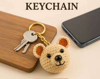May include: A crocheted bear keychain with brown ears and a black nose. The keychain is attached to a gold key ring and chain. The bear is on a wooden surface next to a black smartphone.