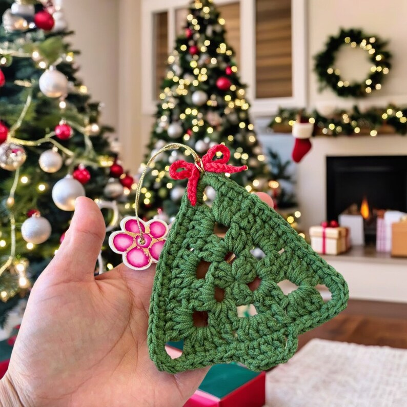 K&ouml;nnte beinhalten: Ein gr&uuml;ner geh&auml;kelter Weihnachtsbaum-Anh&auml;nger mit einer rosa Blumenverzierung und einer roten Schleife. Der Anh&auml;nger wird in einer Hand vor dem Hintergrund eines geschm&uuml;ckten Weihnachtsbaums gehalten.