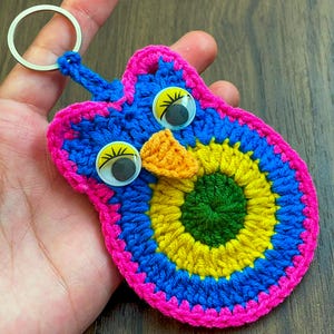 May include: A colourful, handmade crochet owl keyring. The owl has a blue body, a yellow and green centre, and a pink border. It features large googly eyes and an orange beak, attached to a silver keyring.