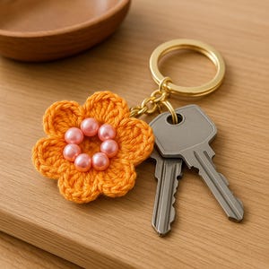 May include: A crocheted orange flower keychain with pink pearl beads. The keychain is attached to a gold key ring and two silver keys.