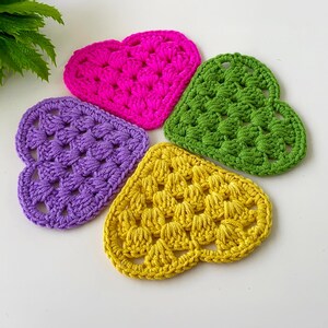 May include: Four heart-shaped crocheted coasters in bright colours: hot pink, green, purple, and yellow. The coasters are made with a textured stitch, perfect for adding a pop of colour to any room. These handmade coasters are ideal for protecting surfaces.