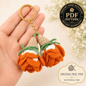 May include: A crochet rose keychain featuring two orange and white flowers with tan centers and green stems attached to a gold-tone metal ring. The image includes text stating Crochet Rose Keychain PDF Pattern Instant Download.