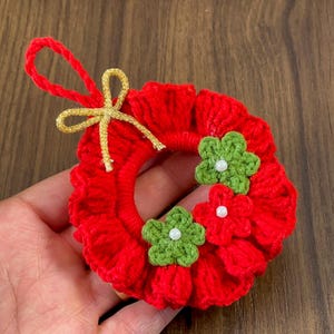 May include: A small red crocheted wreath with a gold ribbon and three green crocheted flowers with white beads in the center.