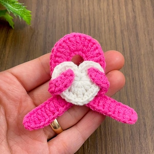 May include: A handmade crochet heart-shaped applique in bright pink and white. The pink crochet forms a ribbon shape, with a white heart in the center. The applique is likely for crafting or embellishment.
