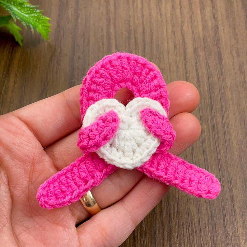 May include: A handmade crochet heart-shaped applique in bright pink and white. The pink crochet forms a ribbon shape, with a white heart in the center. The applique is likely for crafting or embellishment.