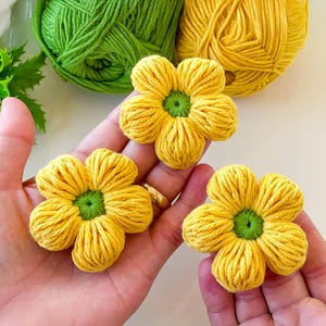Puede incluir: Tres flores amarillas de ganchillo hechas a mano con centros verdes se sostienen en las manos de una persona. Dos ovillos de hilo, uno verde y otro amarillo, están en el fondo. Las flores tienen cinco pétalos cada una y están hechas de hilo.