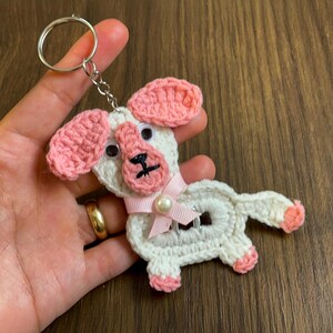 Könnte beinhalten: Weißer gehäkelter Hundefiguren-Schlüsselanhänger mit rosa Ohren, einer rosa Nase und einer rosa Schleife mit einer Perle. Der Schlüsselanhänger ist an einem silbernen Schlüsselring befestigt.