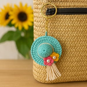 May include: A teal crocheted hat keychain with a white brim and a pink and yellow flower embellishment. The keychain is attached to a gold chain and a woven bag.