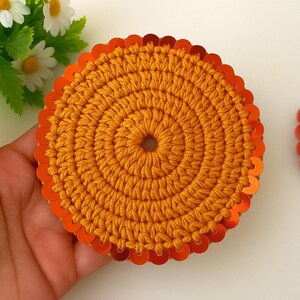 May include: A round crocheted coaster with a bright orange color and a border of orange sequins.