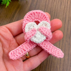 May include: A handmade crochet applique in the shape of a pink ribbon with a white heart in the center. The ribbon is held in a hand, showcasing the delicate stitching and texture of the yarn. The applique is a charming craft item.