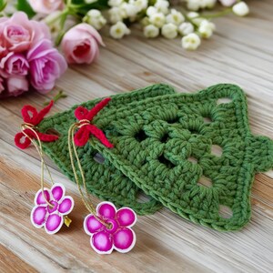 K&ouml;nnte beinhalten: Zwei gr&uuml;ne, geh&auml;kelte Weihnachtsbaum-Anh&auml;nger mit roten und goldenen Schleifen. Die Anh&auml;nger sind mit rosa Blumen verziert.