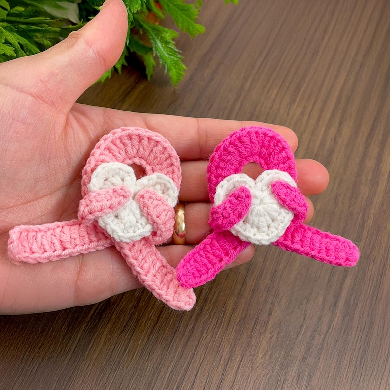 May include: Two handmade crochet appliques in shades of pink and white. Each applique features a circular element with a heart-shaped design in the center and a long, ribbon-like extension. The appliques are held in a hand.